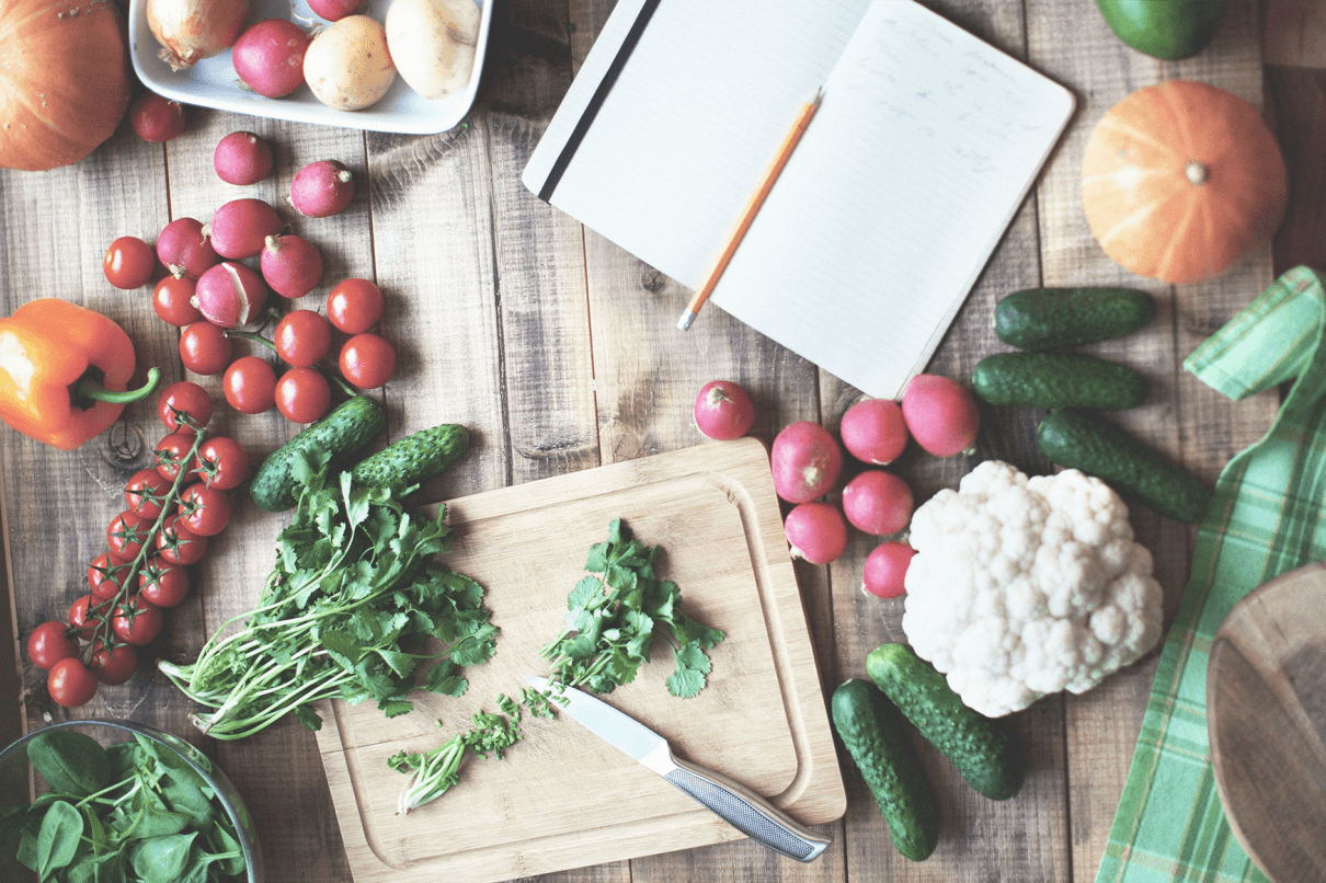 Overhead view of colorful, fresh vegetables, herbs, and an open notebook on a wooden table. Use these among our tips for sharing recipes!