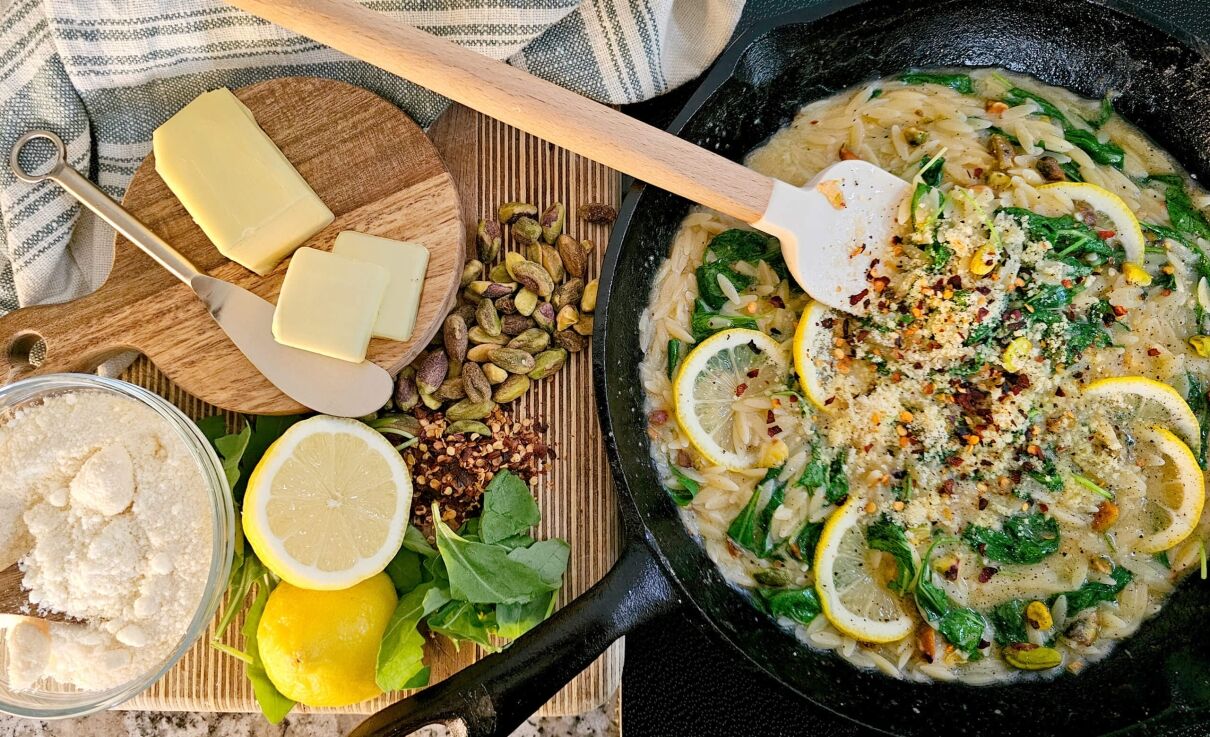 GRO-Lemon Orzo Sept Blog Banner A cast-iron skillet filled with creamy lemon orzo pasta, garnished with spinach, lemon slices, pistachios, red pepper flakes, and parmesan. A spatula rests in the skillet. To the side, a wooden board holds butter slices, whole pistachios, red pepper flakes, fresh lemons, spinach leaves, and a bowl of grated parmesan cheese. A striped kitchen towel is draped nearby.