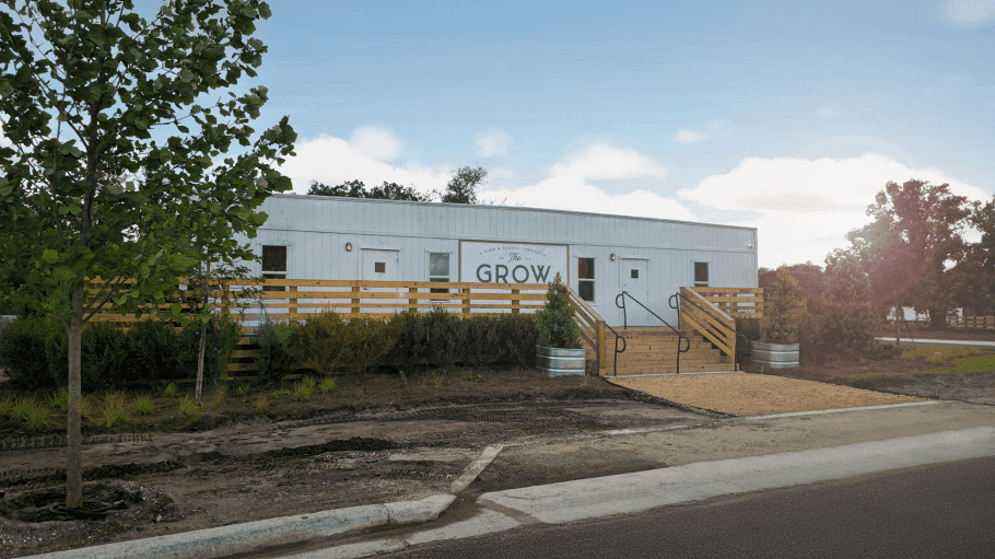 Exterior view of welcome center where new home sales underway at The Grow — a white farmhouse-style building with wooden stairs, ramp access, landscaped greenery, and “The Grow” signage on the front under a bright blue sky.