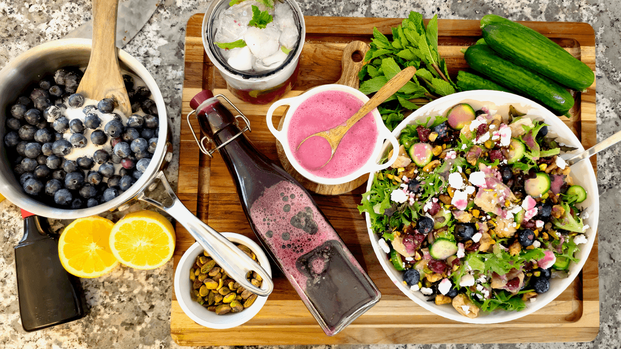 A wooden board displays fresh ingredients and a finished blueberry salad: a saucepan of blueberries being stirred with a wooden spoon, a bottle and small bowl of bright pink blueberry dressing, a glass of iced mint water, sliced lemons, pistachios, fresh mint, cucumbers, and a large bowl of mixed greens topped with blueberries, cucumbers, nuts, and crumbled cheese.