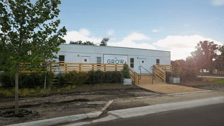 Exterior view of welcome center where new home sales underway at The Grow — a white farmhouse-style building with wooden stairs, ramp access, landscaped greenery, and “The Grow” signage on the front under a bright blue sky.