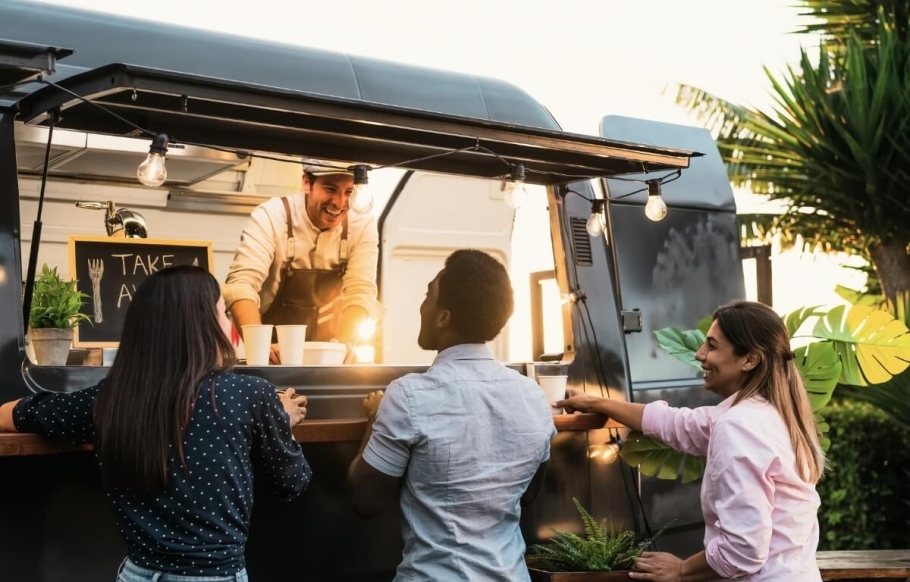 A group of people gather at a food truck, where a smiling person is ready to help them. They are surrounded by trees and greenery.