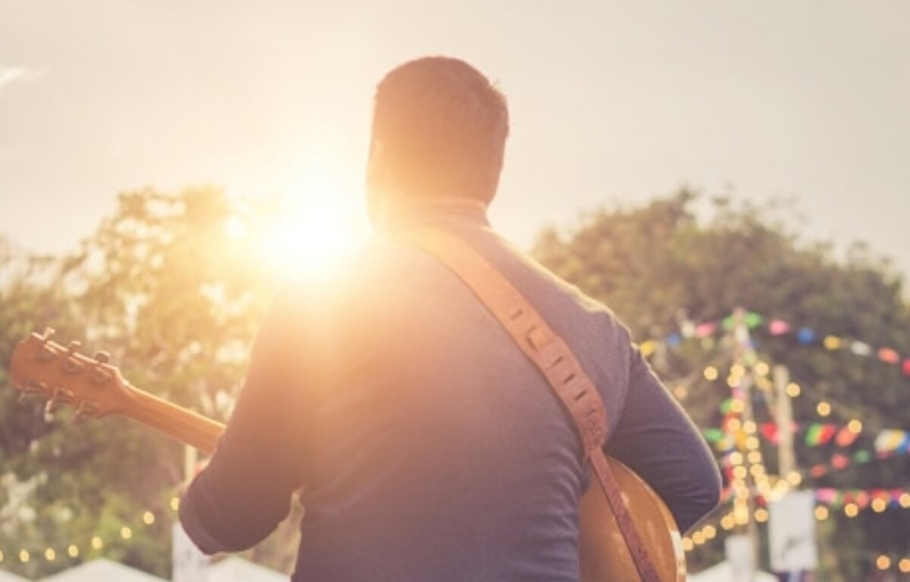 An image showing the back of a person playing an acoustic guitar. The sun is beginning to set, and fairy lights surround the stage — part of the lifestyle at The Grow.