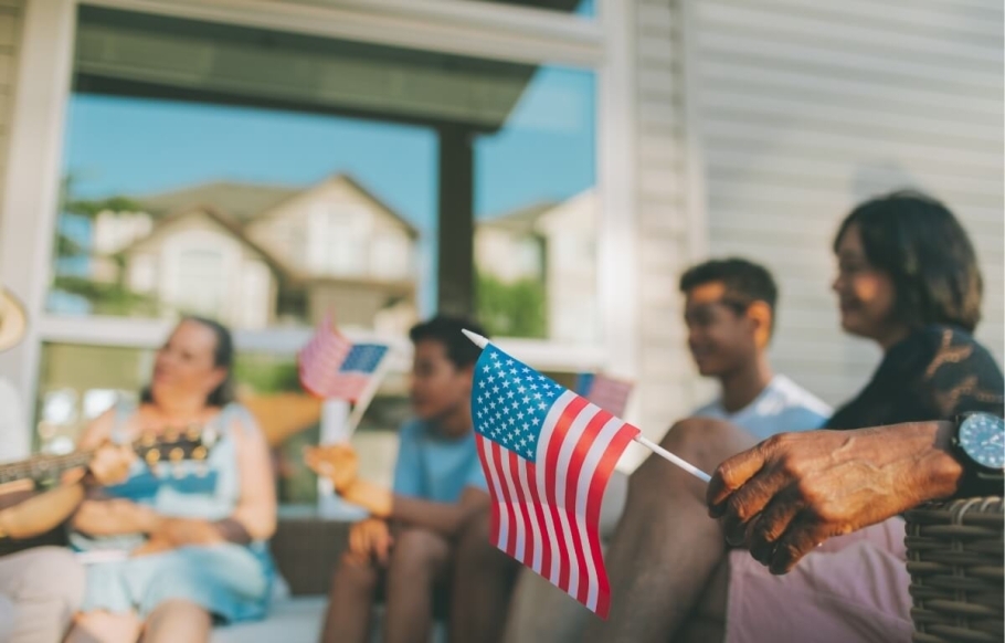 A group of people in soft focus on a front porch or patio are sitting around listening to a guitar player on the left. In the foreground on the right is a hand holding a small American flag in sharp focus — part of the lifestyle at The Grow.