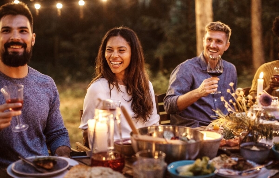 A group of people holding wine glasses aloft in a toast are sitting around a full outdoor table at night under twinkling cafe lights.