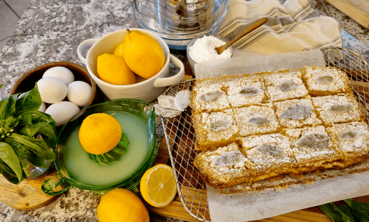 GRO-lemon-basil-bars An overhead view of fresh baked lemon basil bars, cut into squares and cooling on a wire rack atop a wooden board. Whole and halved lemons, eggs, confectioner's sugar, basil leaves, and a juicer with lemon juice are artfully arranged next to the bars, on a stone countertop.